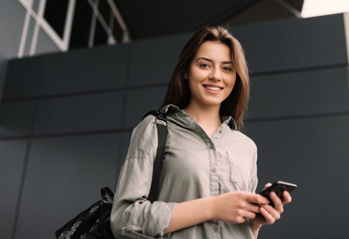 Portrait of positive and smiling university student using mobile phone for researching information online. Attractive successful woman walking on street and posing for pictures. Online education.